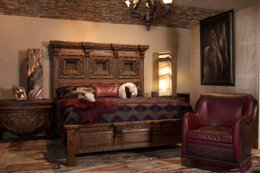 Western-style bedroom with hand-carved wooden bed, red leather chair, patterned bedding, and warm ambient lighting in a rustic stone-accented lodge setting.