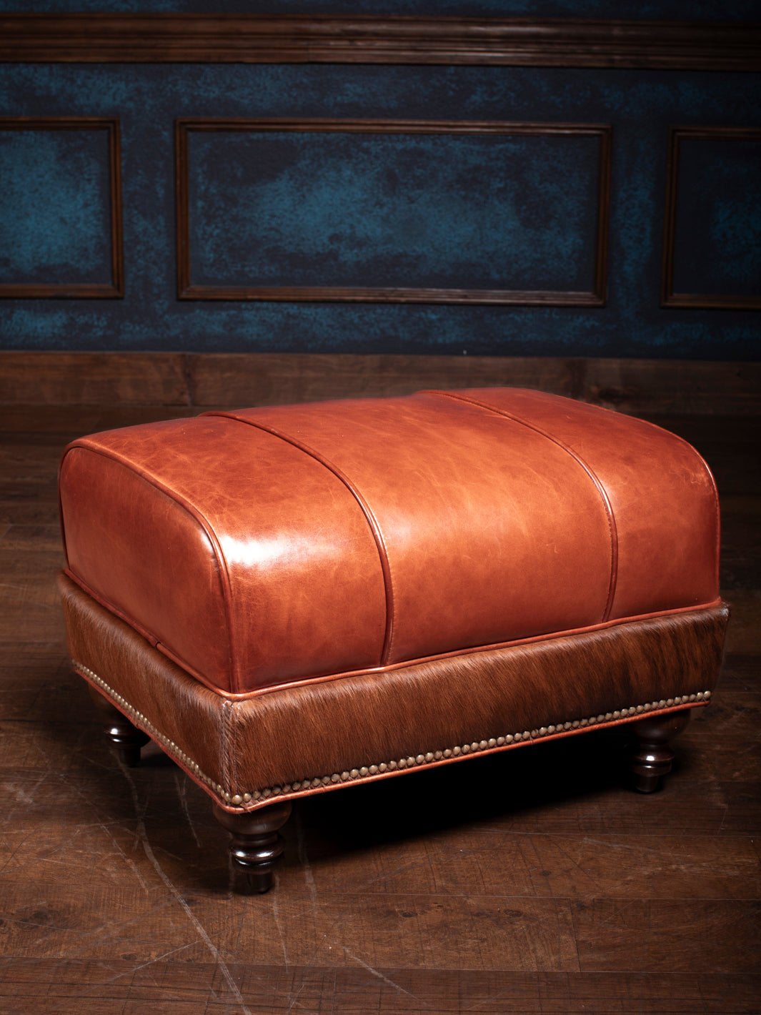 Amarillo Red Stitch Ottoman with rich leather top and hair-on-hide bottom, featuring brass nailhead trim and sturdy wood legs.
