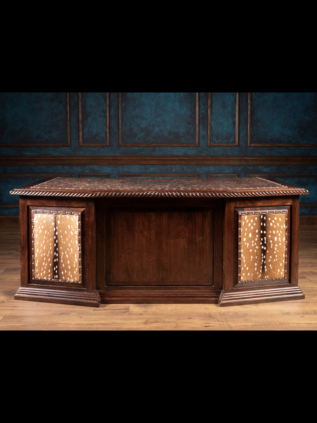 A wooden executive desk with a leather surface, metal studded nailhead detailing, and an axis hide panel accent, set against a blue panelled wall.