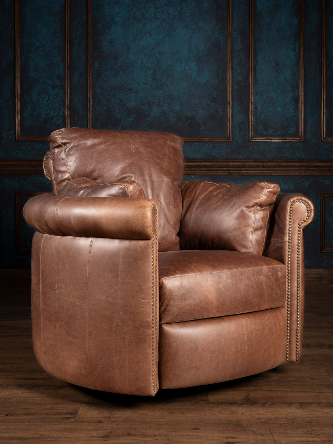 A brown leather snug swivel recliner chair with rolled arms and nailhead trim, placed against a blue panelled wall.