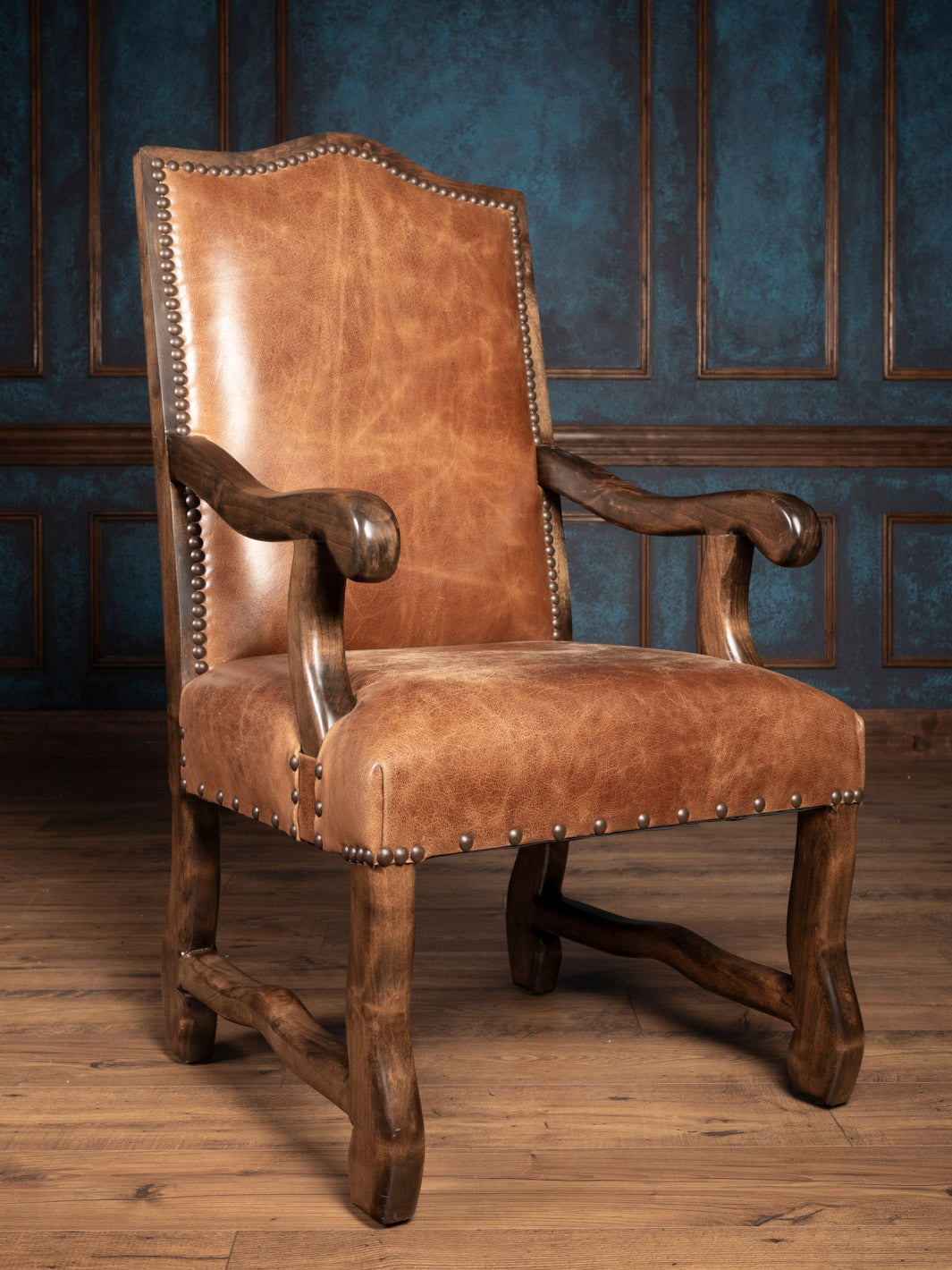 A brown buffalo leather dining arm chair with a solid wood frame and antique nailhead trim, displayed against an indoor backdrop.