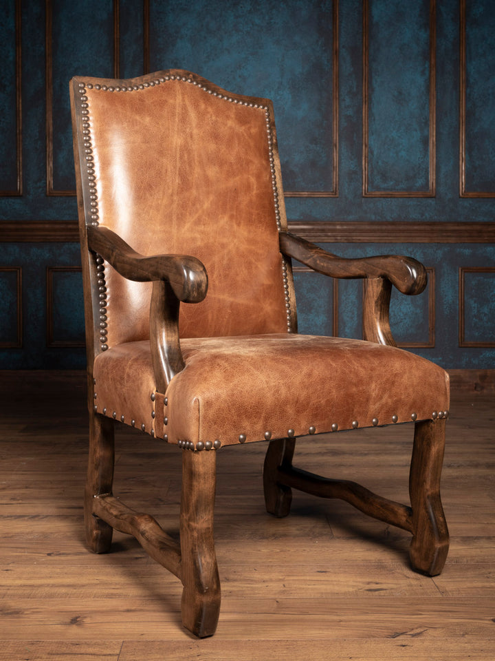 A brown buffalo leather dining arm chair with a solid wood frame and antique nailhead trim, displayed against an indoor backdrop.