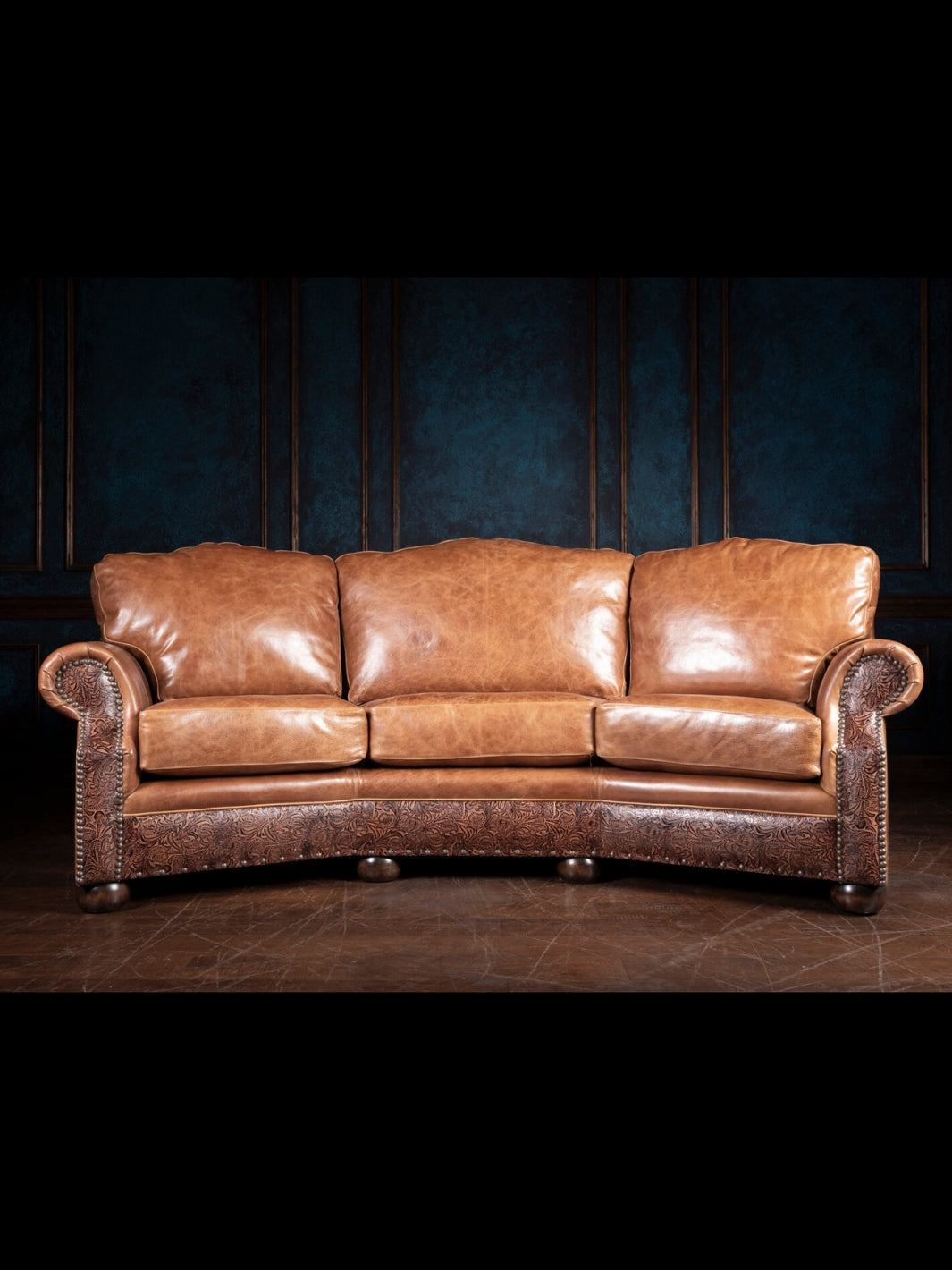 A brown leather curved sofa with nailhead trim and hand-tooled detailing, set against a dark paneled wall.