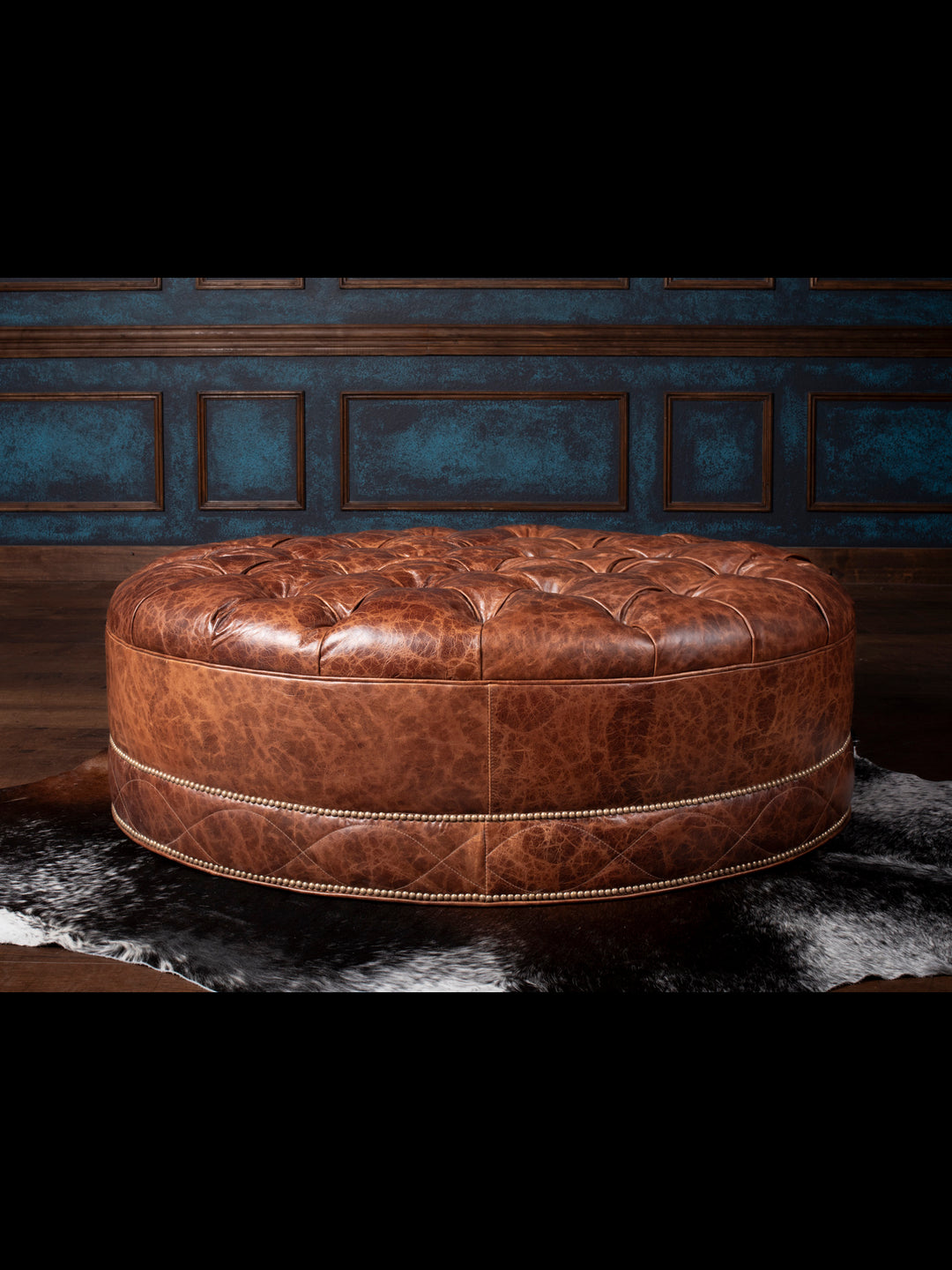 An oval-shaped tufted ottoman with caramel-colored leather and nailhead trim, placed on a dark floor with a blue wall in the background.