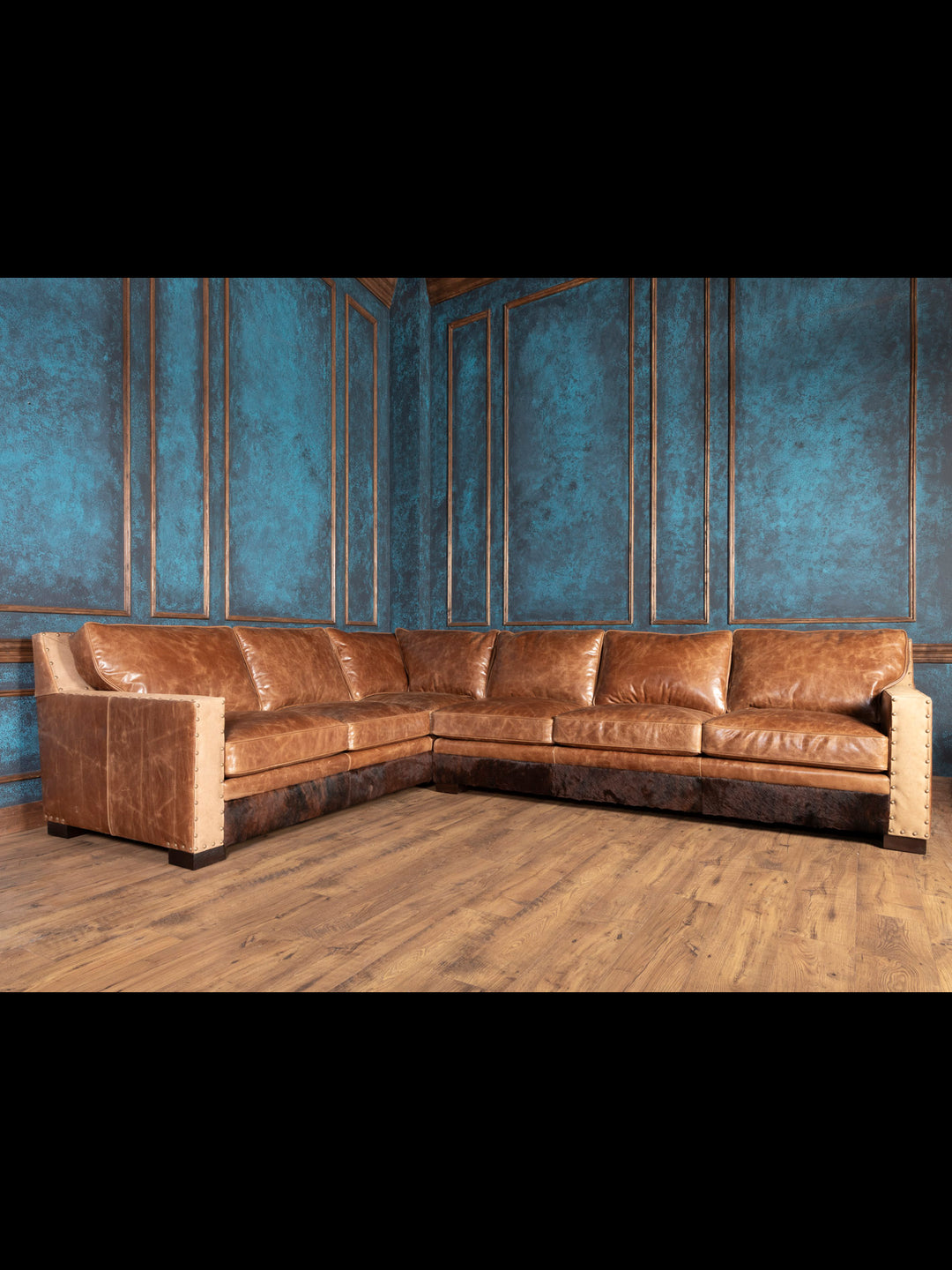 A large, L-shaped brown leather sectional sofa with cowhide accents, placed in a room with blue walls and wooden flooring.