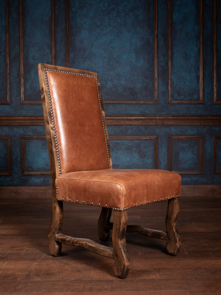 A brown leather dining chair with a high back and nailhead trim, featuring a wooden frame, positioned against a blue panelled wall.