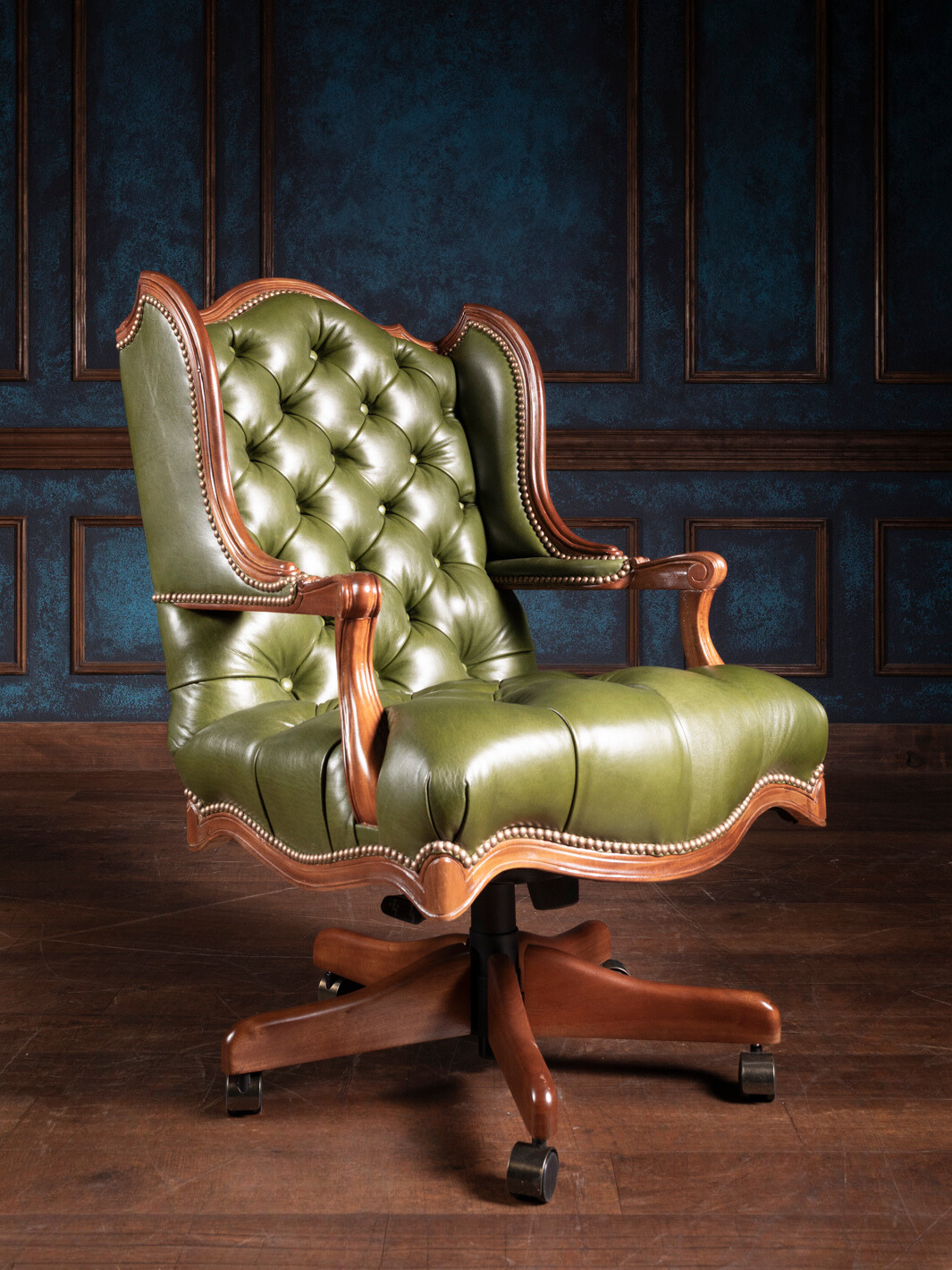 A green tufted leather desk chair with classic tufting and nailhead detailing, on a wooden floor with a blue paneled wall in the background.