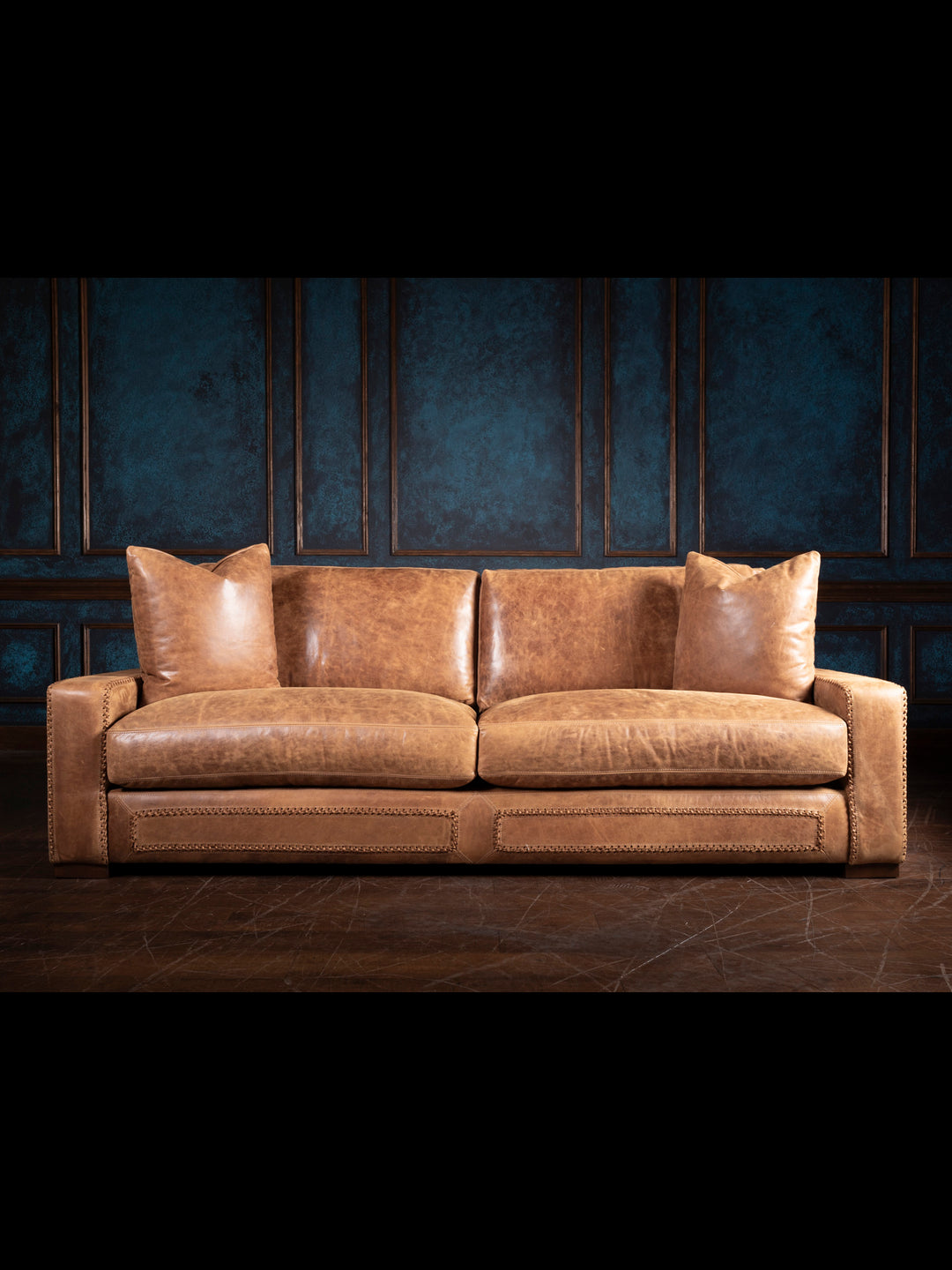 Full view of a rustic tan leather sofa with deep cushions and wide armrests, styled in a dark wood-paneled room.