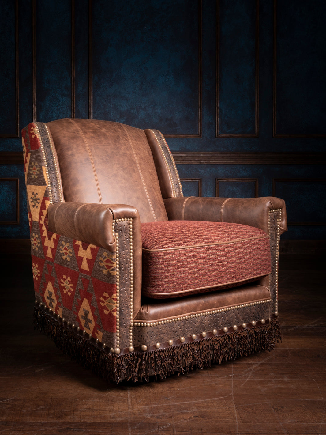 A swivel chair with a combination of leather and fabric upholstery, featuring a rich brown leather back, a bold red fabric seat, and a decorative fringe detail at the base, set against a dark blue background.