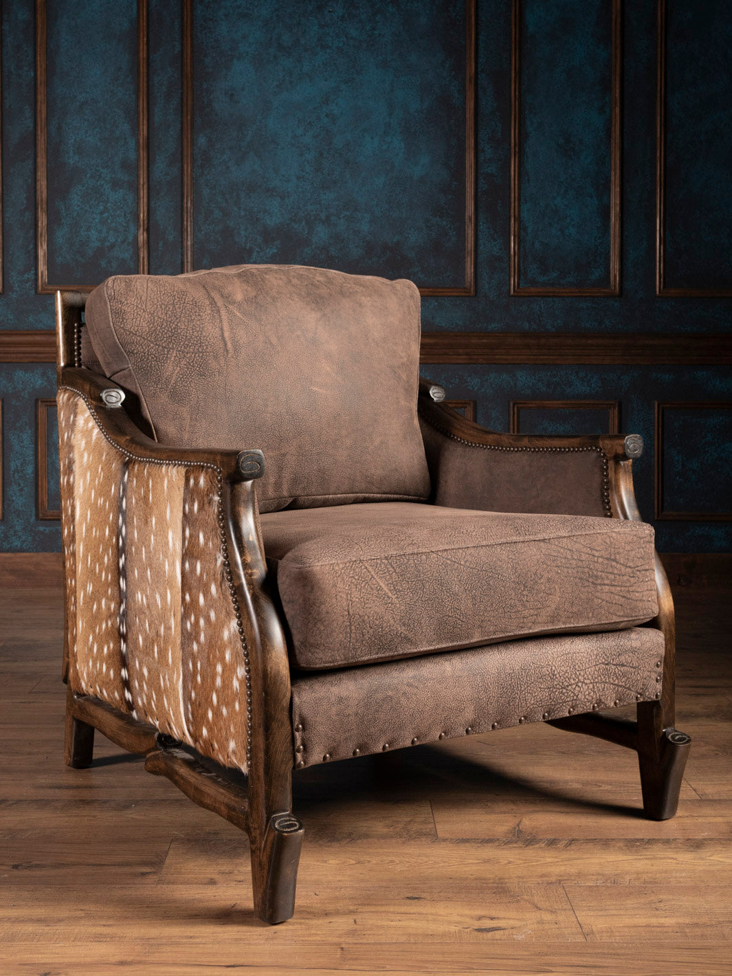 A brown leather accent chair with axis deer hide accents on the armrests and backrest, placed against a blue textured background.