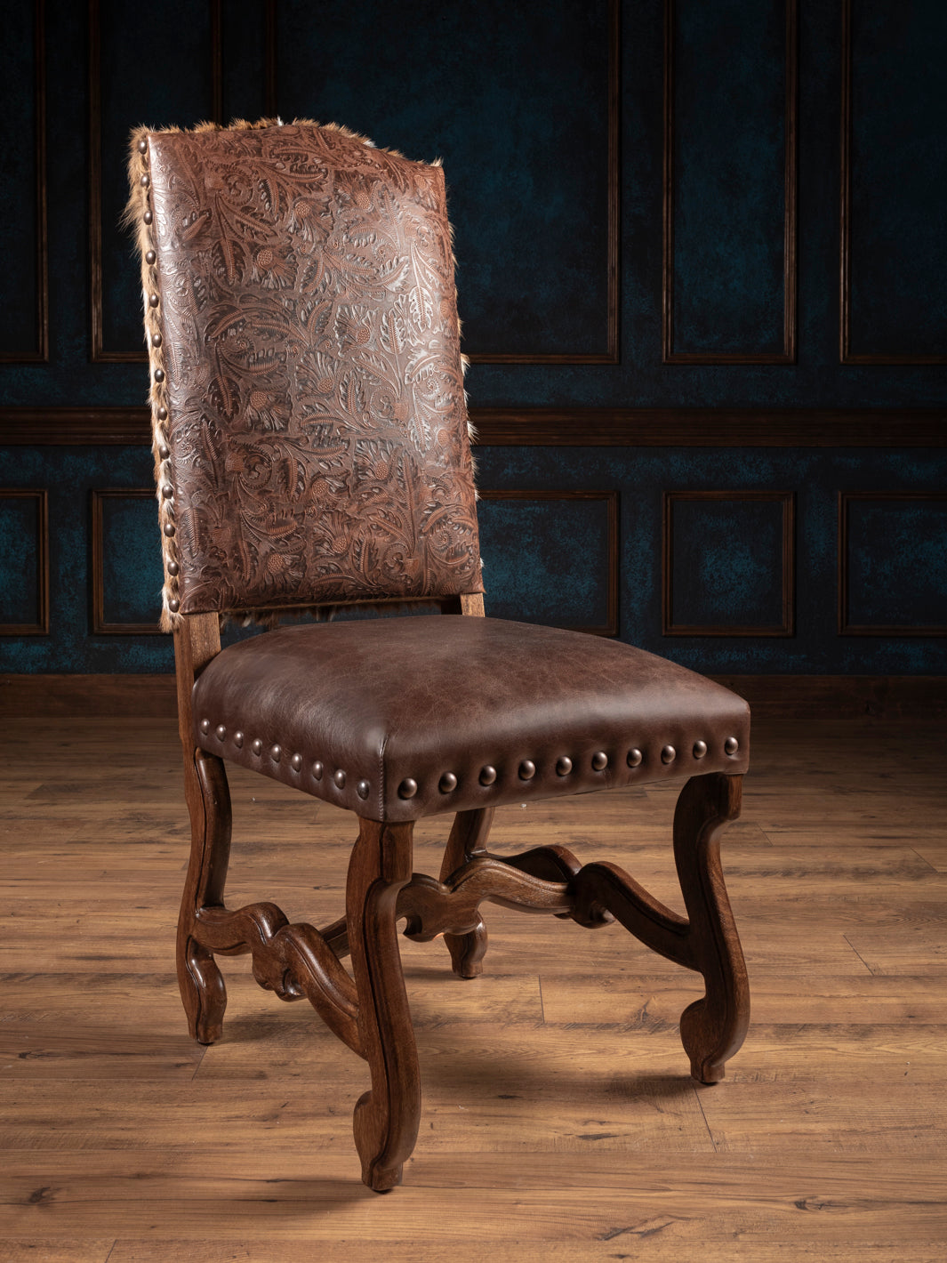 A brown leather dining chair with a hand-carved wooden frame and upholstered backrest, displayed against a dimly lit panelled wall.
