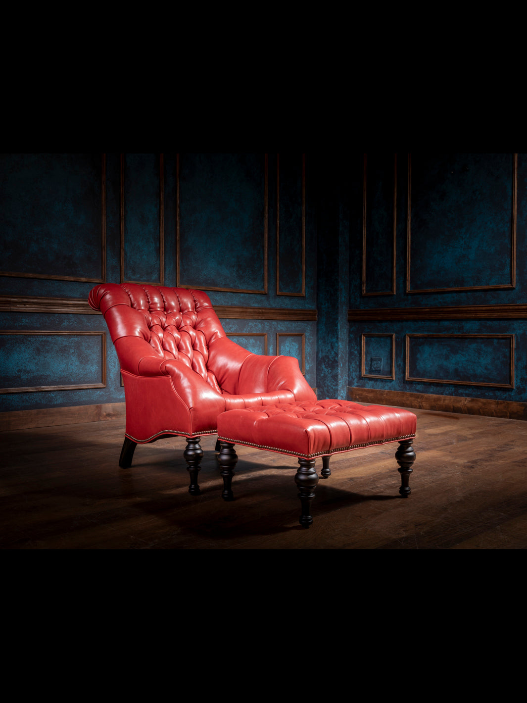 Tufted red leather accent chair with ottoman, featuring a handcrafted silhouette, rolled back, and Western-style detailing in a moody blue-paneled room.
