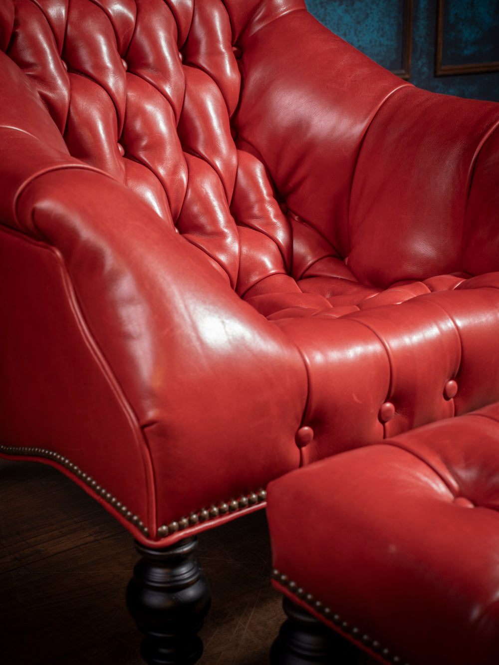 Detail view of a tufted red leather accent chair with plush cushioning, sculpted arms, and antique brass nailhead trim, paired with a matching ottoman.