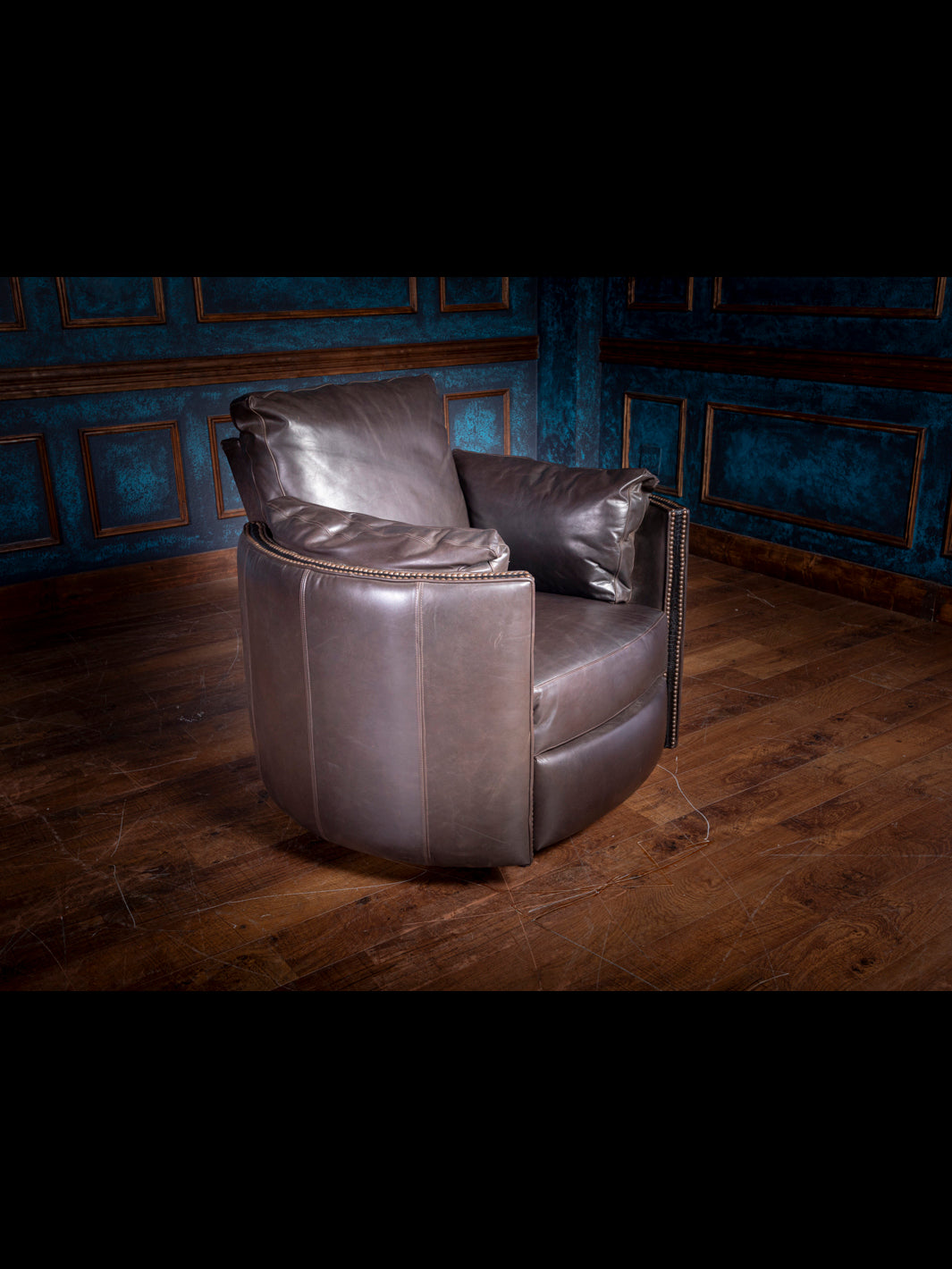 A charcoal gray leather snug swivel recliner chair positioned on a wooden floor against an old blue paneled wall.