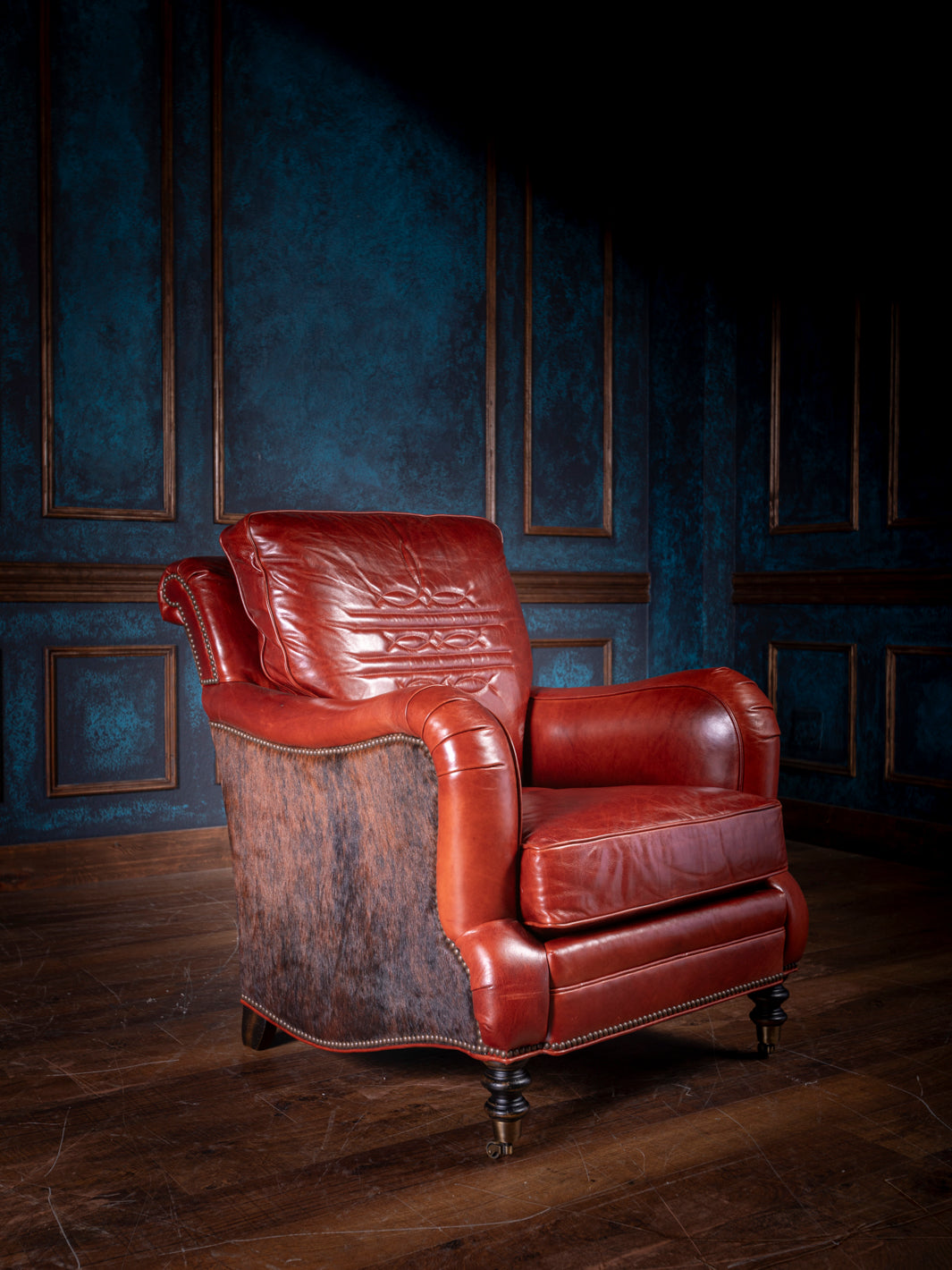 A western luxury leather chair featuring rich red leather, rolled arms, and a cowhide back panel, set against a dark wood and navy backdrop. The nailhead trim and turned wooden legs enhance its upscale, rustic charm.