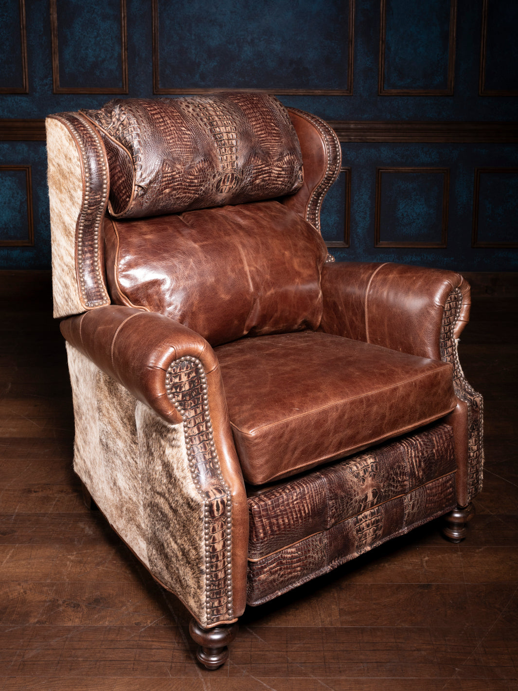 A croc-embossed leather recliner chair with brown cushions and natural cowhide accents on the arms and backrest, placed against a blue panelled wall.
