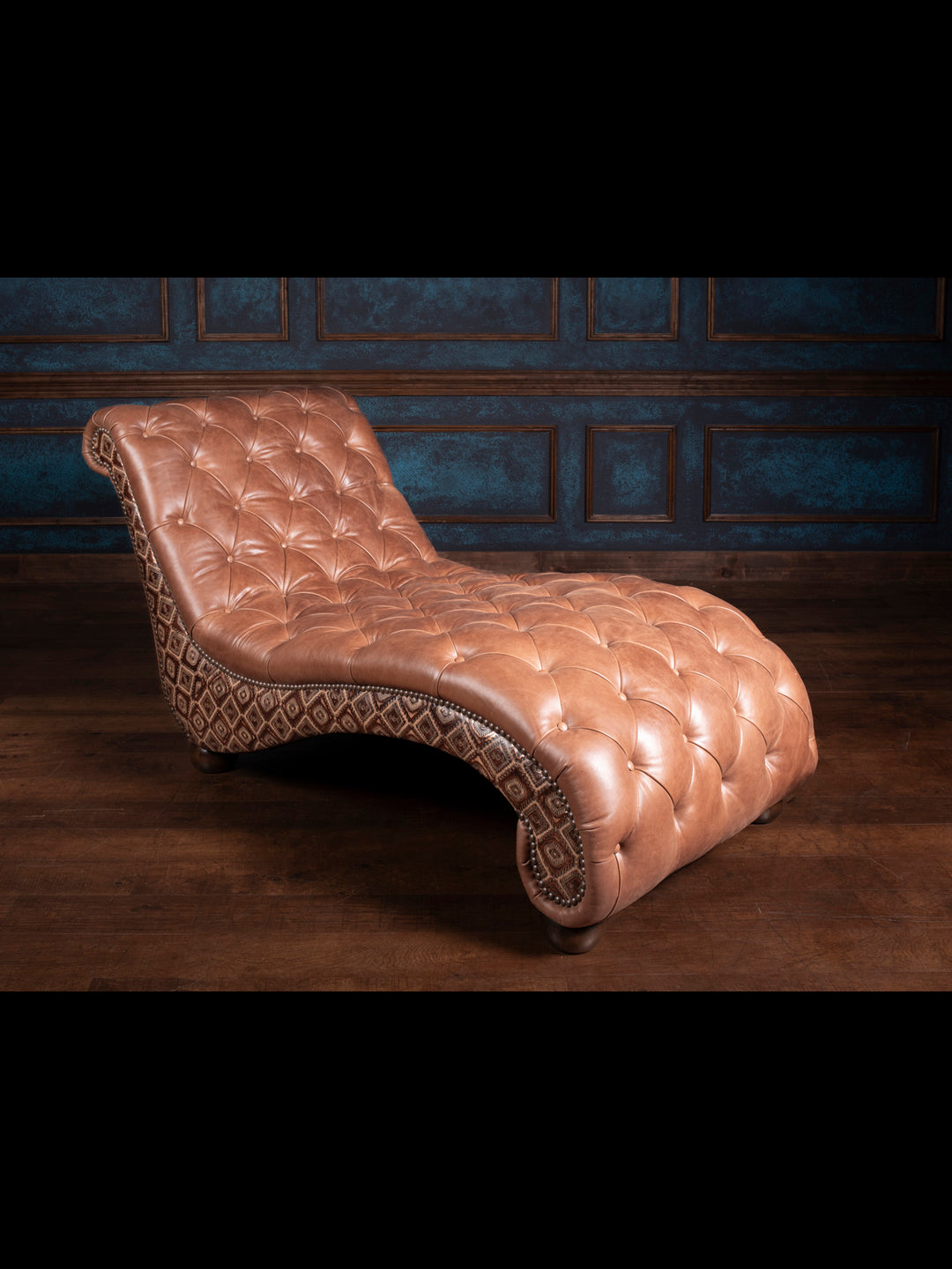 A rustic mahogany leather chaise lounge with tufted seats and embossed artisan inlays, placed in a room with a dark blue wall and wooden floor.