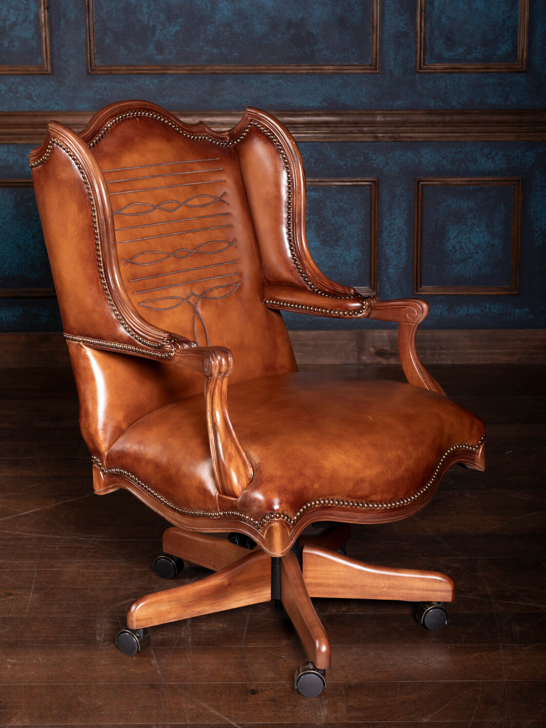 A brown leather desk chair with saddle stitching, featuring an ergonomic design, on a wooden floor against a blue panelled wall.