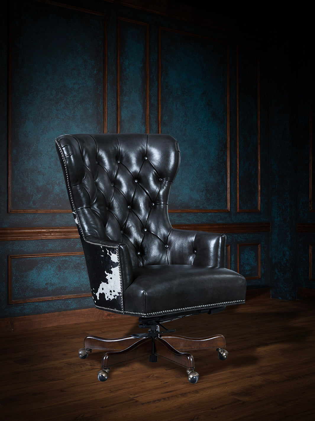 A black speckled cowhide leather wingback desk chair with a rich leather seat and button-tufted backrest, on a wooden floor with blue panelled walls in the background.
