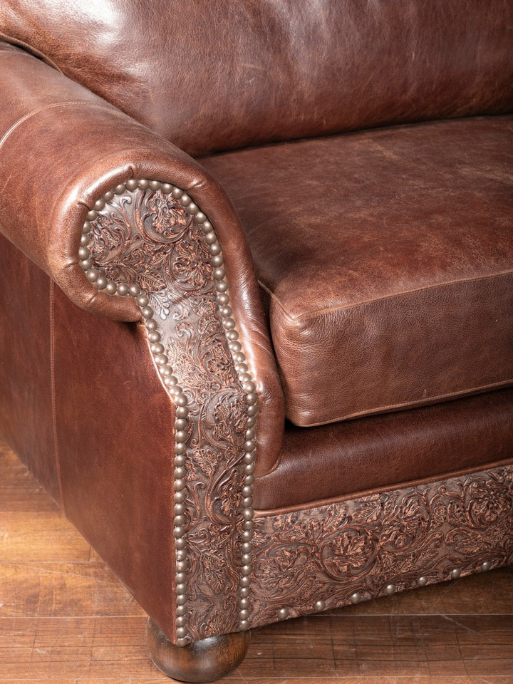 Detailed view of tooled leather on the arm of a Western-style oversized chair, accented with antique brass nailheads and smooth full-grain leather upholstery.