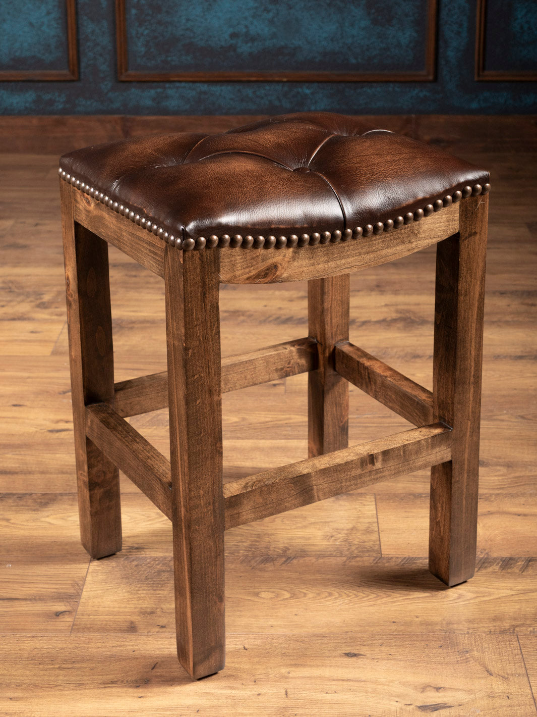 A brown tufted leather saddle stool with a solid wood frame, featuring a tufted backless seat and a nailhead trim.