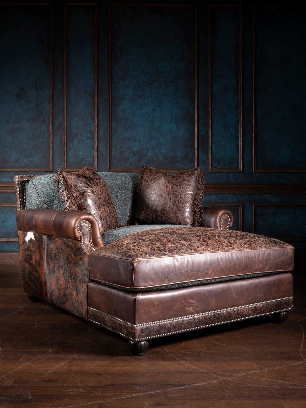 A turquoise leather and fabric chaise lounge with dark leather accents and patterned fabric cushion, placed against a blue paneled wall.