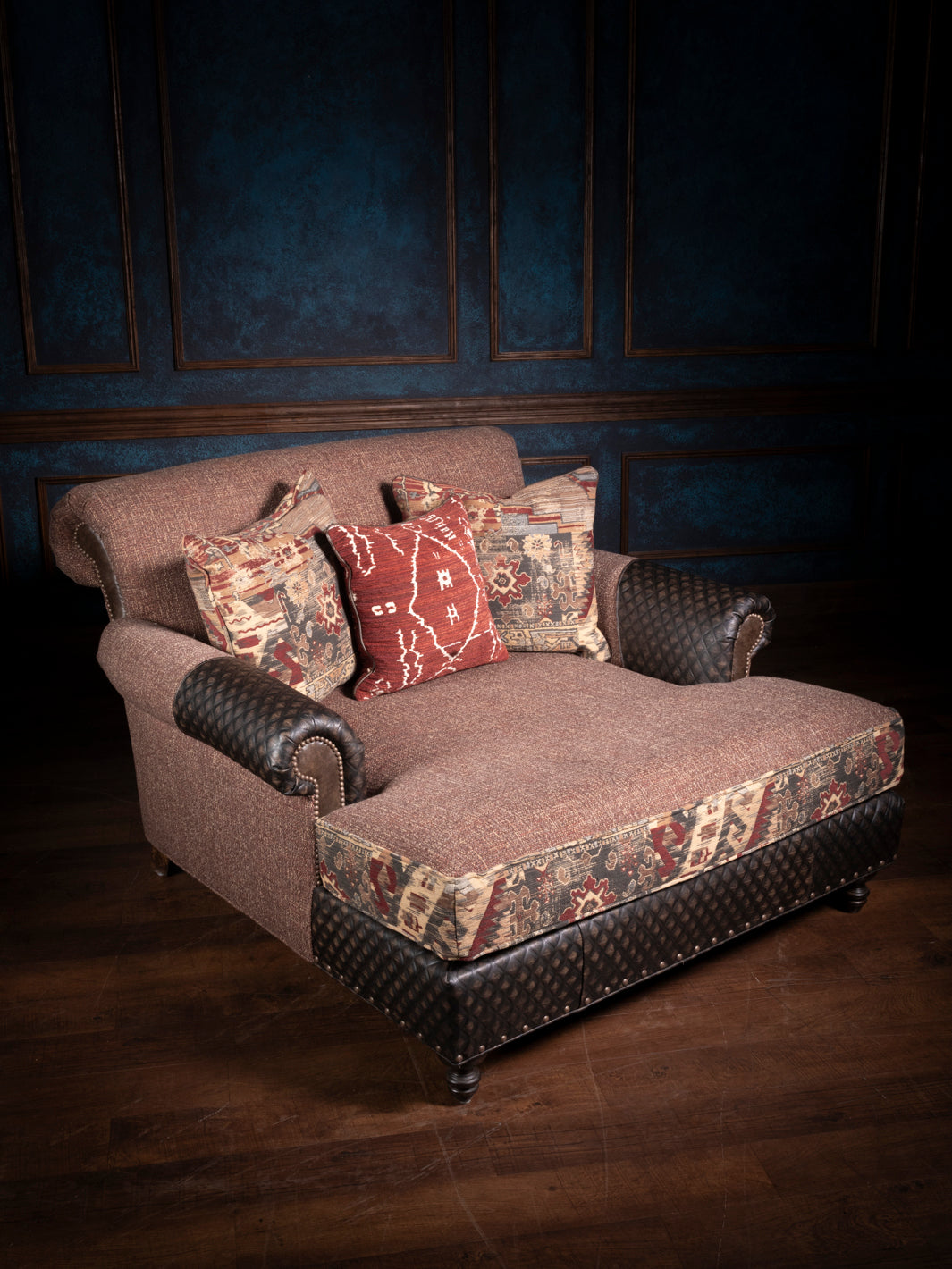A chaise lounge chair with a combination of brown leather and patterned fabric, featuring plush cushions and a durable structure, set against a blue panelled wall.