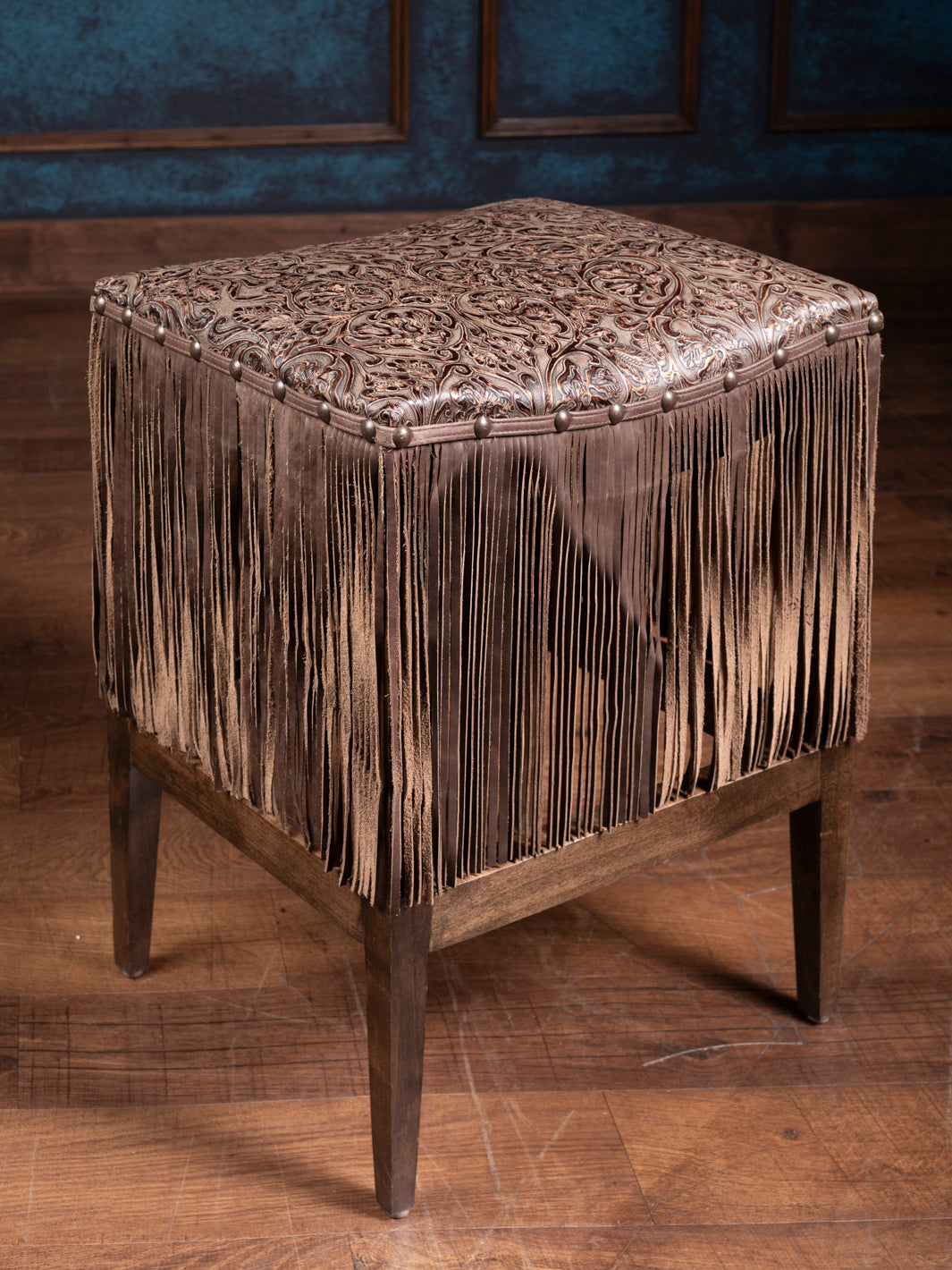 A brown leather saddle stool with fringe detailing and embossed patterns.