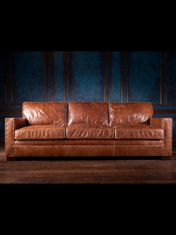 A brown, full-grain leather sofa with a wax finish, displayed in a room with dark walls.