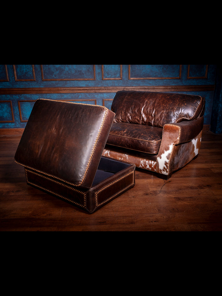 Functional leather ottoman with hidden storage, shown open in front of a hair-on-hide Western lounge chair with distressed brown leather upholstery.
