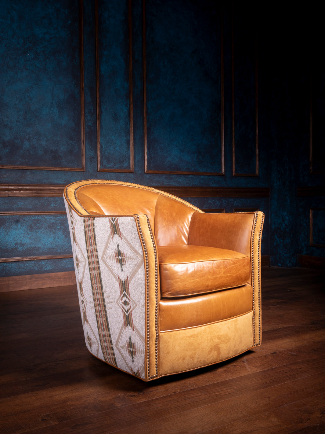 A brown leather swivel chair with adobe-style fabric accents and suede leather trim, placed against a blue wall on a wooden floor.