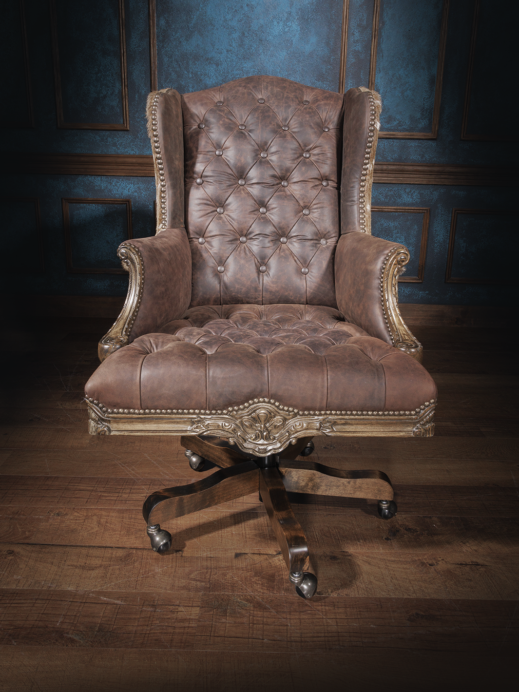 Front view of an office chair with button-tufted brown leather upholstery, wingback design, nailhead trim, and a carved wooden swivel base with casters.