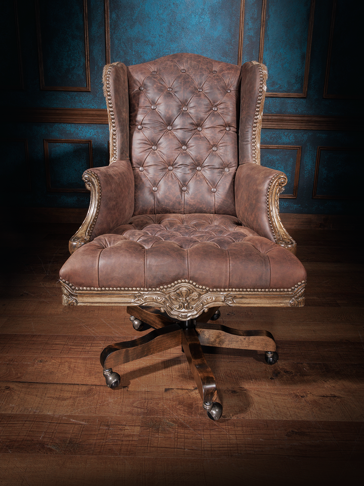 Front view of an office chair with button-tufted brown leather upholstery, wingback design, nailhead trim, and a carved wooden swivel base with casters.