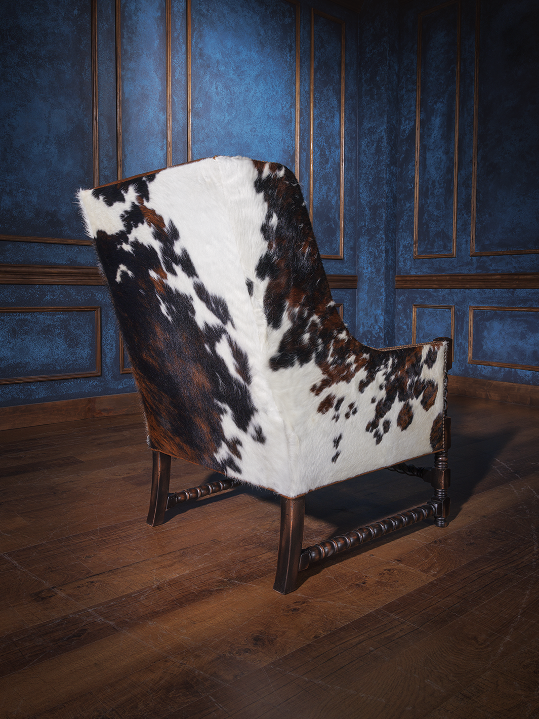 Rear view of the western-style armchair fully wrapped in tri-color cowhide, showing off its bold hide pattern and gently curved silhouette over a wooden floor.