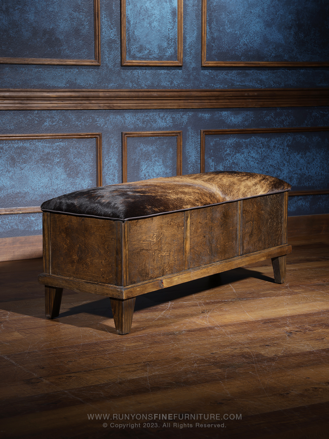 A Western cowhide trunk with dark hair on a brown wooden surface, placed against a blue paneled wall. The trunk is closed and used as a bench.