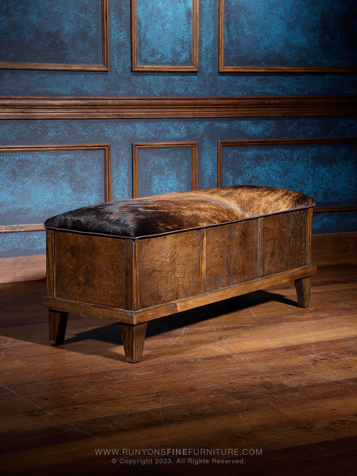 A Western cowhide trunk with dark hair on a brown wooden surface, placed against a blue paneled wall. The trunk is closed and used as a bench.