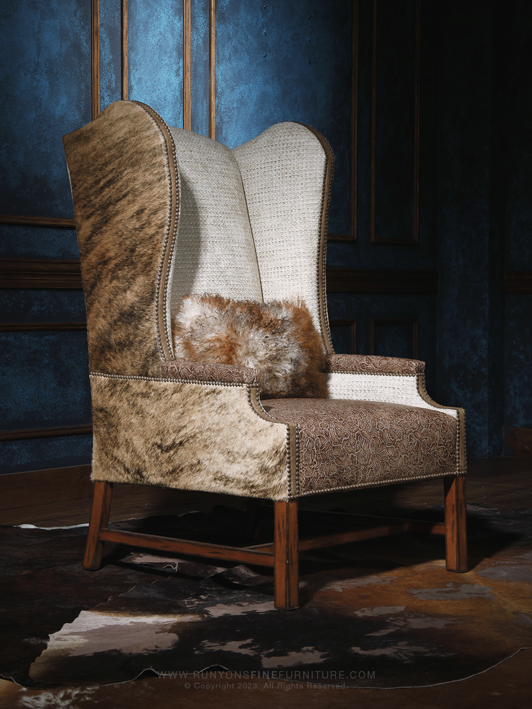 Western cowhide wingback chair featuring a tall sculpted back with a blend of soft beige fabric and natural hide sides. The brown tooled leather seat and decorative nailhead trim give it a refined rustic elegance, accented by a plush fur pillow.