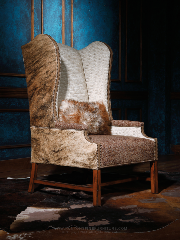 Western cowhide wingback chair featuring a tall sculpted back with a blend of soft beige fabric and natural hide sides. The brown tooled leather seat and decorative nailhead trim give it a refined rustic elegance, accented by a plush fur pillow.
