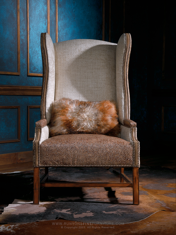 Front view of a western cowhide wingback chair showcasing a high textured fabric back, ornate brown upholstery on the seat, and a coordinating fur accent pillow. Nailhead trim outlines the curves, enhancing its luxury-meets-frontier look.
