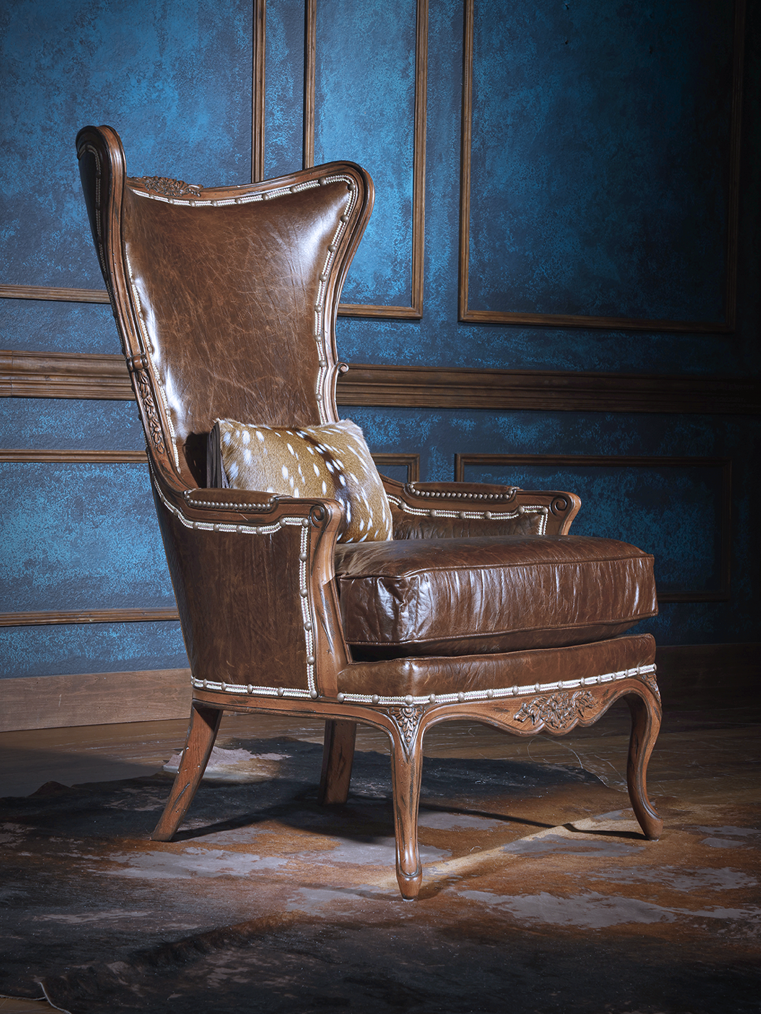 Side angle of a high-back leather accent chair with sculpted wood frame, smooth leather panels, and a western-style pillow in spotted axis hide. The chair exudes classic lodge sophistication.