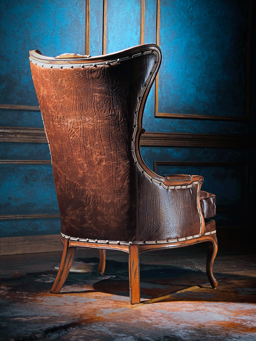 Back view of a western leather wingback chair showcasing its tall, textured leather back and hand-carved wood base. A fusion of stately craftsmanship and rugged elegance.