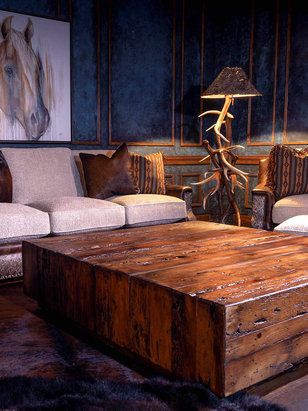 view of wooden coffee table in the center sitting on hair on hide rug, surrounded by leather and fabric sofa and matching leather and fabric accent chair next to antler lamp, with a horse painting on the blue wall in the back
