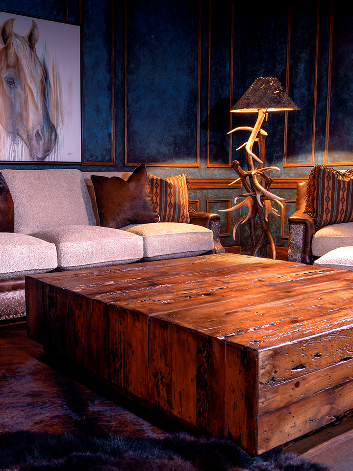 view of wooden coffee table in the center sitting on hair on hide rug, surrounded by leather and fabric sofa and matching leather and fabric accent chair next to antler lamp, with a horse painting on the blue wall in the back