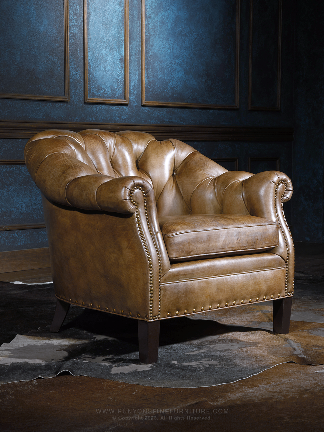 Western leather Chesterfield chair in a warm saddle brown finish, featuring deep tufted upholstery, rolled arms, and nailhead trim. The chair sits on tapered wooden legs over a hide rug in a richly styled interior