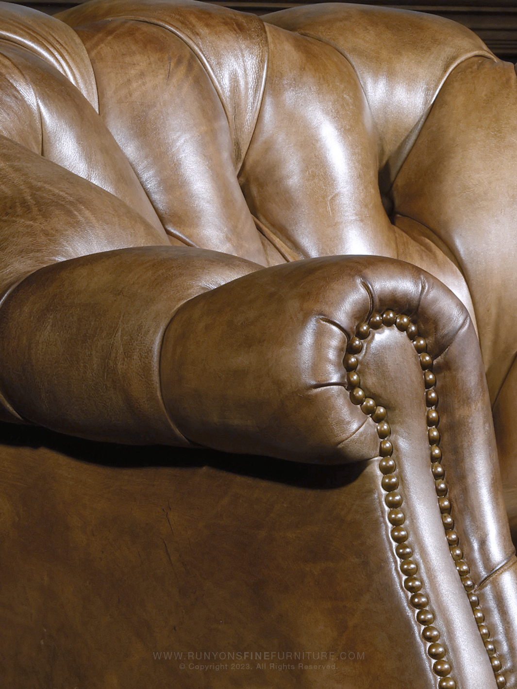 Close-up of a tufted leather arm and rolled backrest, highlighting the supple texture and detailed brass nailhead trim of this western-style Chesterfield accent chair.