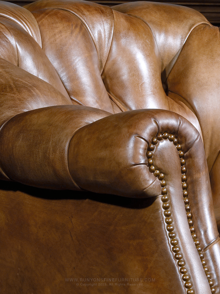 Close-up of a tufted leather arm and rolled backrest, highlighting the supple texture and detailed brass nailhead trim of this western-style Chesterfield accent chair.