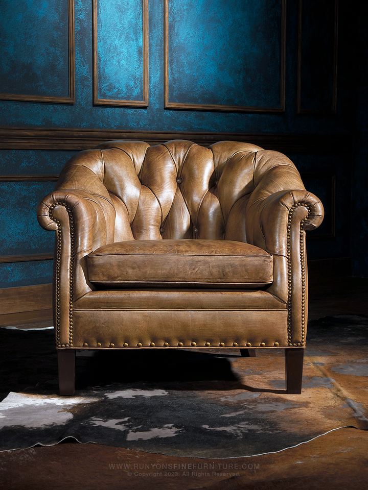 Front-facing western Chesterfield chair with deep channel tufting, plush seat cushion, and bold rolled arms. The aged saddle leather finish and brass nailheads deliver timeless rustic elegance.