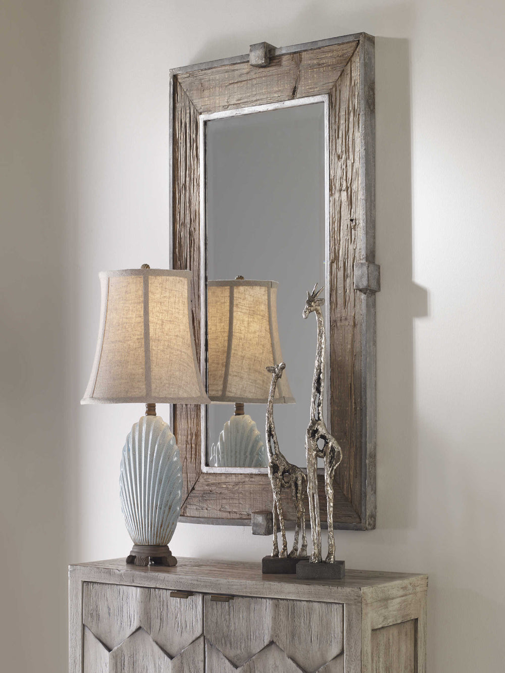 Decorative table with a lamp, shell sculpture, and giraffe statue in front of a wooden-framed mirror.