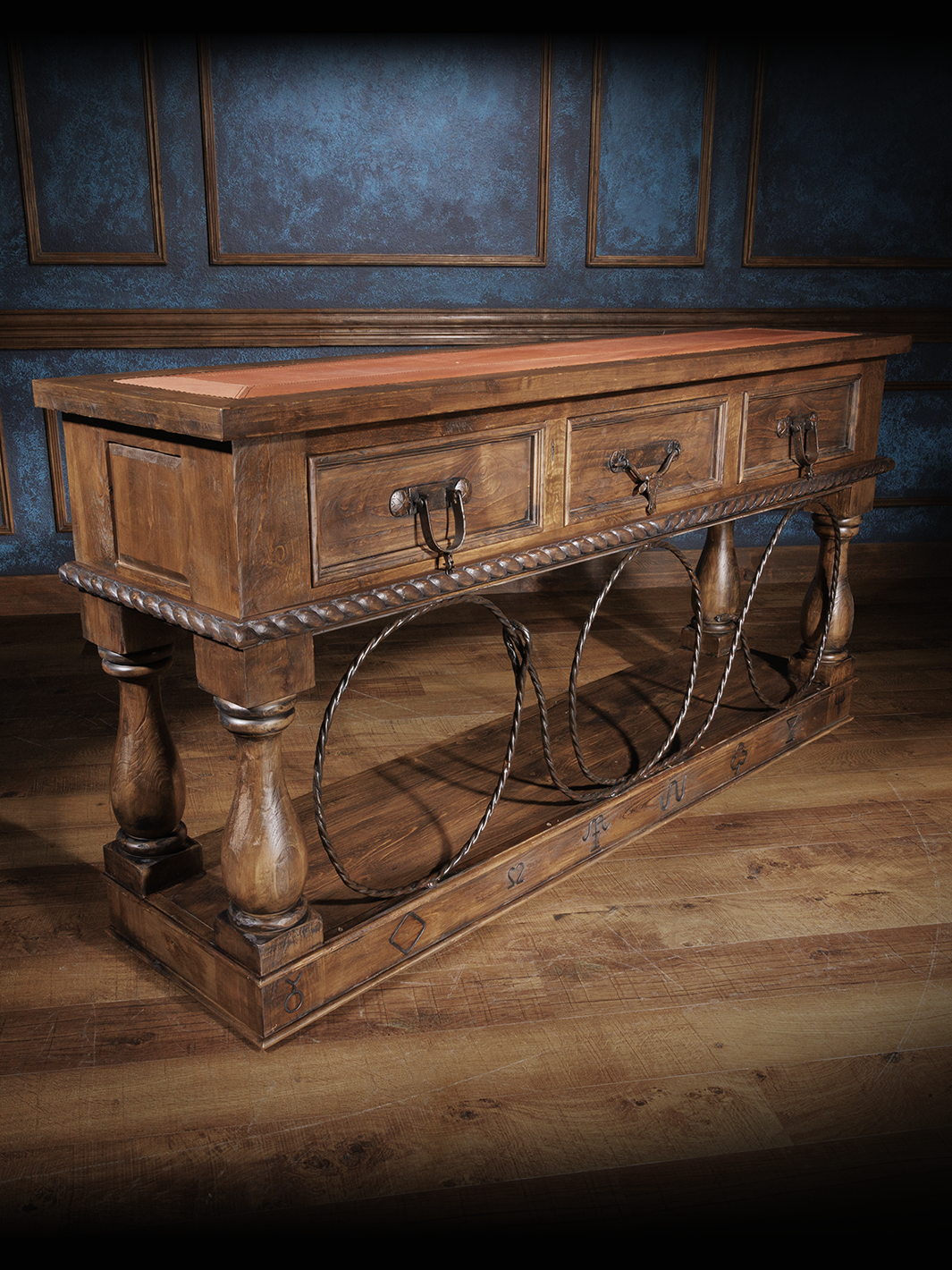 Rustic wooden console table with a brown leather inlay, metal ring accents, and ornate carvings, set against a dark wall background.