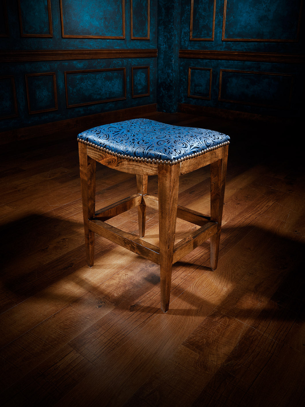 A wooden western saddle stool with a blue upholstered seat, positioned on a hardwood floor against a paneled wall.